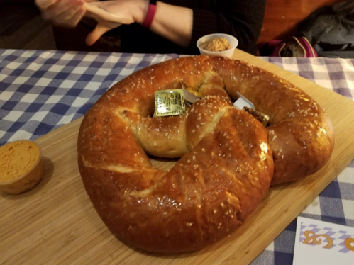 A large, soft pretzel on a wooden board, accompanied by butter and a small cup of dipping sauce.
