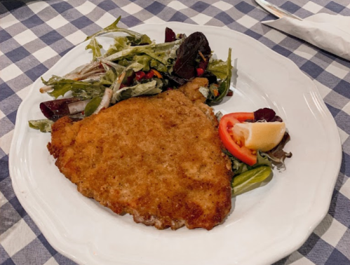 A breaded fish fillet served with a side salad and a slice of lemon on a white plate, set on a checkered tablecloth.