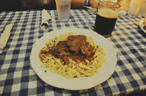 A plate of beef stew served over noodles, accompanied by a glass of dark beer, on a checkered tablecloth.