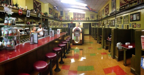 A vintage diner interior with a red counter, bar stools, jukebox, and colorful tiled floor, adorned with memorabilia.
