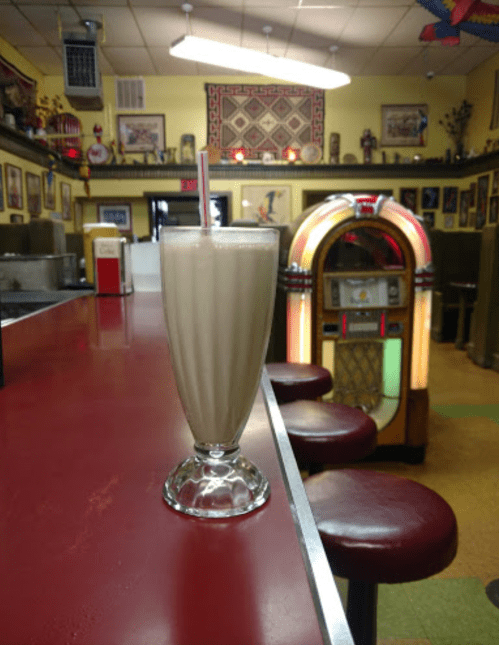 A tall glass of milkshake with a straw sits on a red countertop in a retro diner, with a jukebox in the background.