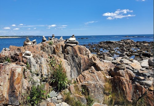A rocky shoreline with stacked stones, blue water, and a clear sky dotted with clouds.