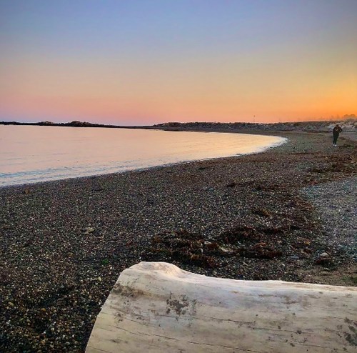 A serene beach at sunset, with calm waters, pebbly shore, and a person walking in the distance.