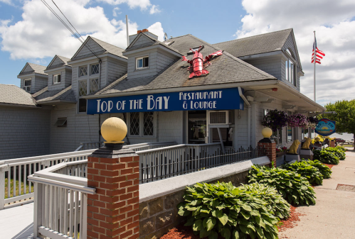 Exterior of Top of the Bay restaurant, featuring a lobster sculpture, flowers, and an American flag on a sunny day.