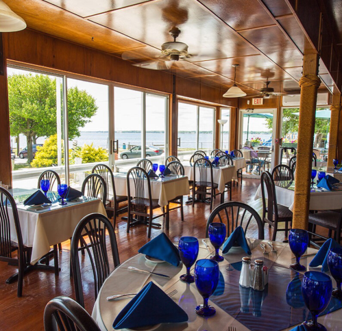 Bright restaurant interior with blue glassware, white tablecloths, and large windows overlooking a scenic waterfront.