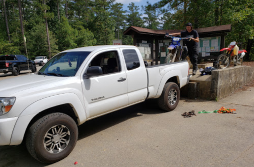 A pickup truck parked near a trailhead, with a person unloading a dirt bike from the truck bed. Trees in the background.
