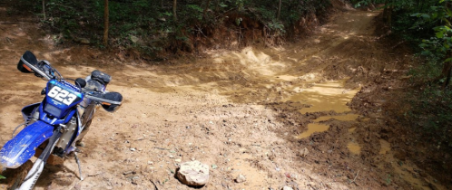 A dirt bike parked beside a muddy trail in a forested area, with a rocky surface and puddles visible.