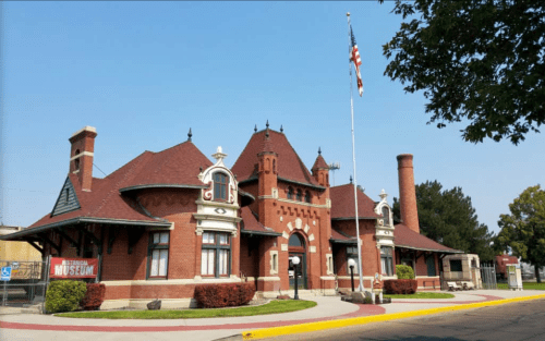 Historic brick train station with a tall chimney, flagpole, and museum sign, set against a clear blue sky.