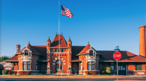 Historic red brick train station with a tall flagpole and American flag, set against a clear blue sky.