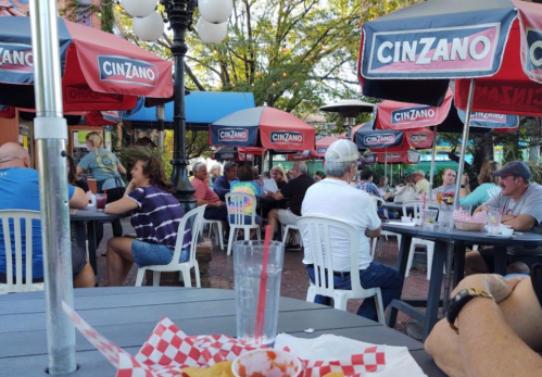 Outdoor dining scene with people at tables under Cinzano umbrellas, enjoying food and drinks in a lively atmosphere.