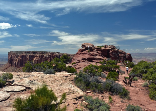 A rocky landscape with green shrubs under a blue sky, featuring a distant cliff and people exploring the area.
