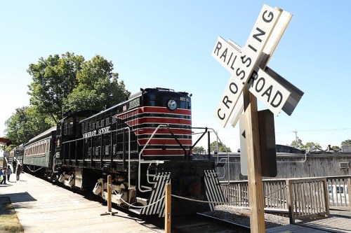 A vintage train at a railroad crossing, surrounded by trees and a wooden platform.