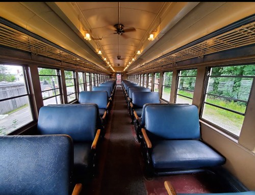 Interior of a vintage train car, featuring rows of blue seats and large windows, with warm lighting and a view outside.