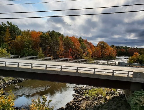 A bridge spans a river surrounded by vibrant autumn foliage under a cloudy sky.