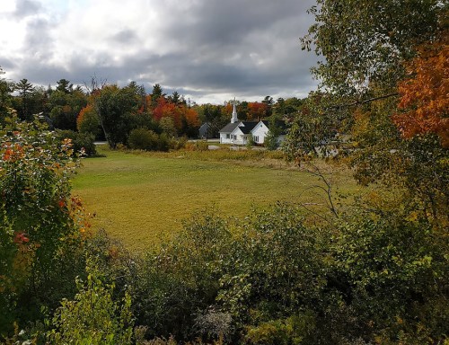 A scenic view of a grassy field surrounded by trees with autumn foliage and a white house in the background.