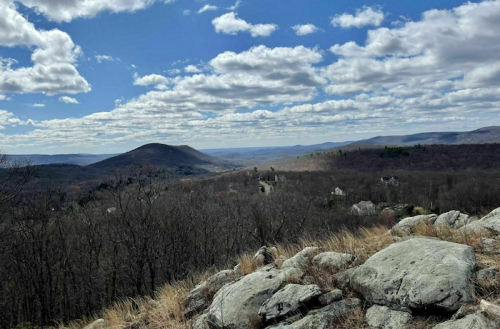 A panoramic view of rolling hills and valleys under a blue sky with fluffy clouds, framed by rocky foreground.