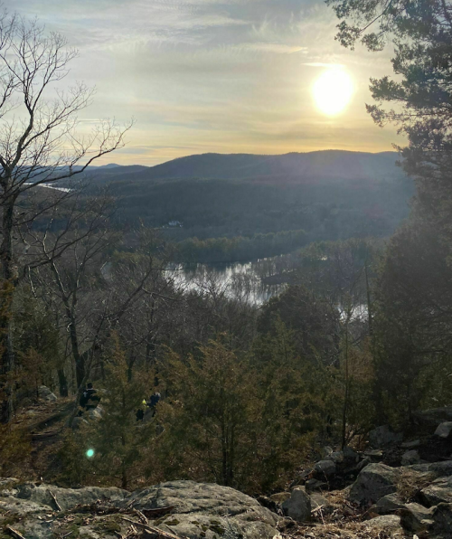 A scenic view of a sunset over rolling hills and a river, framed by trees and rocky terrain.