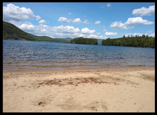 A sandy beach by a calm lake, surrounded by green trees and mountains under a blue sky with fluffy white clouds.