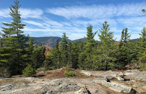 A scenic view of a rocky landscape surrounded by green trees and distant mountains under a blue sky with clouds.