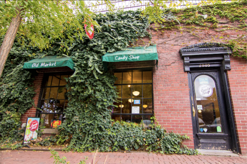A brick building with green awnings for "Old Market" and "Candy Shop," surrounded by lush ivy and a cobblestone path.
