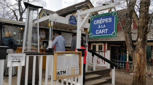 A small outdoor food stand with a sign reading "Crêpes à la Cart," featuring customers in line and nearby trees.