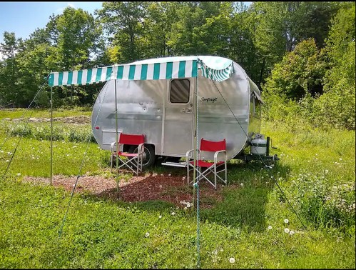A silver vintage trailer with a striped awning, surrounded by green grass and trees, with two red chairs outside.