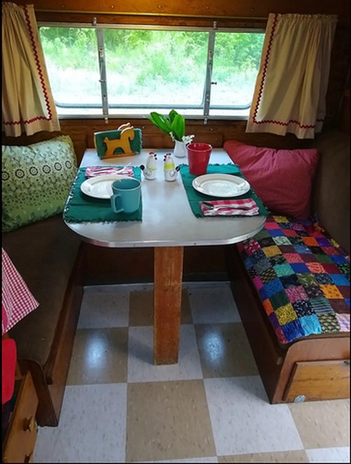Cozy camper interior with a table set for two, colorful cushions, and a window view of greenery outside.