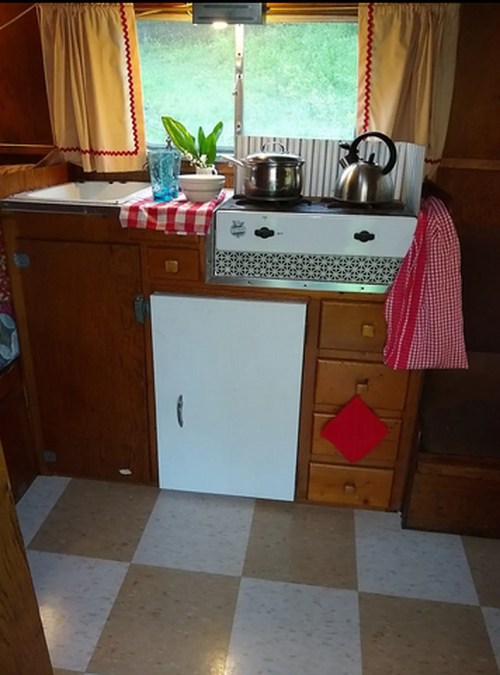 Cozy vintage kitchen with wooden cabinets, a kettle, potted plant, and checkered tablecloth on the counter.