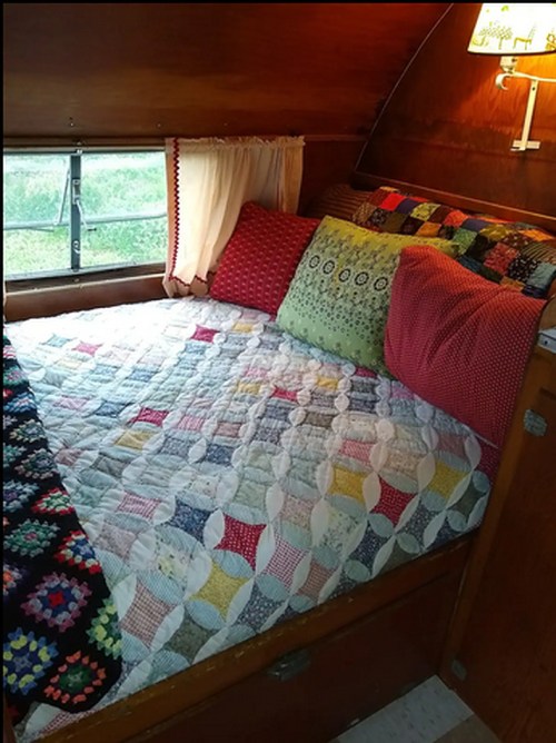Cozy vintage camper interior with a quilted bed, colorful pillows, and a warm lamp by the window.