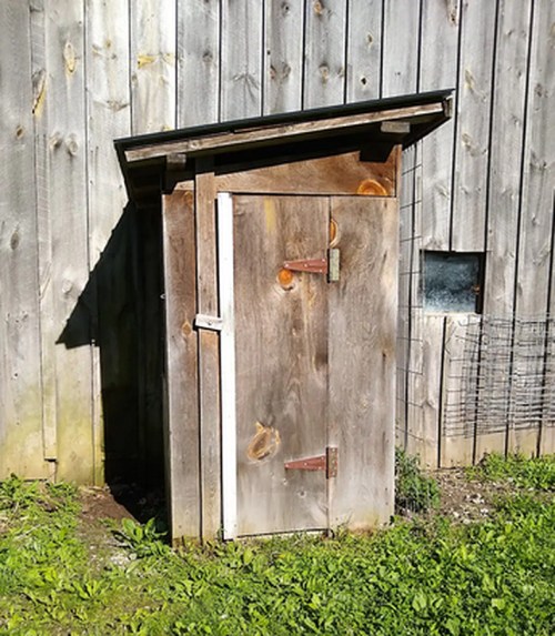A small wooden shed with a slanted roof, standing next to a wooden wall and surrounded by grass.