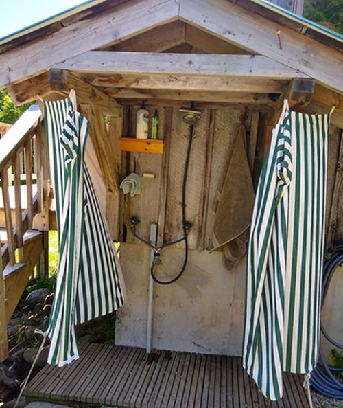Outdoor shower with striped curtains, wooden structure, and various shower fixtures. Surrounded by greenery.