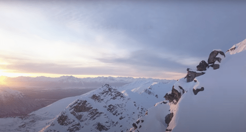 A panoramic view of snow-covered mountains under a soft sunset sky, showcasing rugged peaks and valleys.