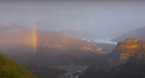 A scenic view of mountains, a glacier, and a rainbow emerging from misty clouds in a serene landscape.