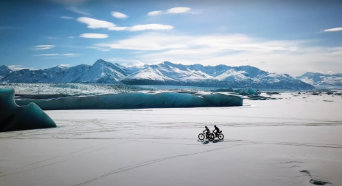 Two cyclists ride fat bikes across a snowy landscape with mountains and ice formations under a clear blue sky.