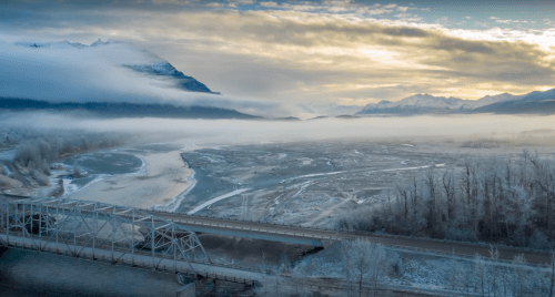 A serene landscape featuring a foggy river, a bridge, and snow-capped mountains under a cloudy sky.