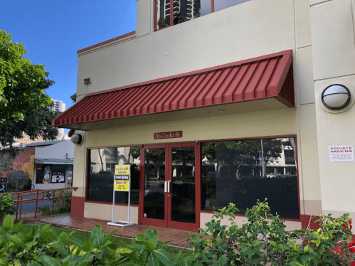 Exterior of a building with a red awning and large windows, featuring a sign for "The Cookie Bar" and nearby greenery.