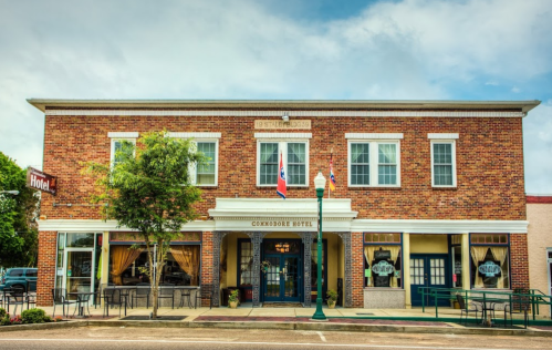 Historic brick building with "Commodore Hotel" sign, featuring large windows and outdoor seating. Flags displayed.