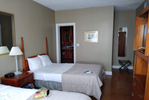 A simple hotel room with two beds, a lamp, a mirror, and wooden furniture against a beige wall.