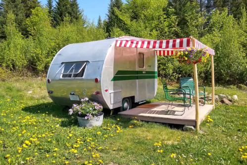 A vintage silver camper with a green stripe, parked under a striped awning, surrounded by flowers and greenery.