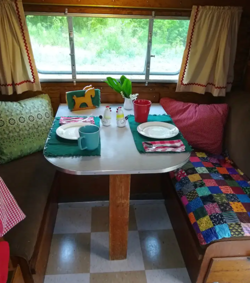 Cozy camper interior with a table set for two, colorful cushions, and a view of greenery outside.