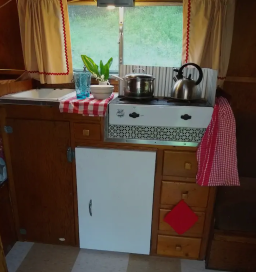 Cozy vintage kitchen with wooden cabinets, a kettle, potted plant, and checkered tablecloth. Bright and inviting atmosphere.