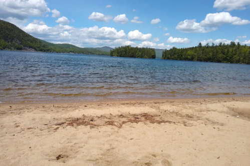 A serene lake view with sandy shore, surrounded by green hills and fluffy clouds in a blue sky.