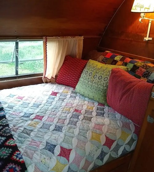 Cozy bedroom with a quilted bedspread, colorful pillows, and wooden walls, featuring a window and warm lighting.