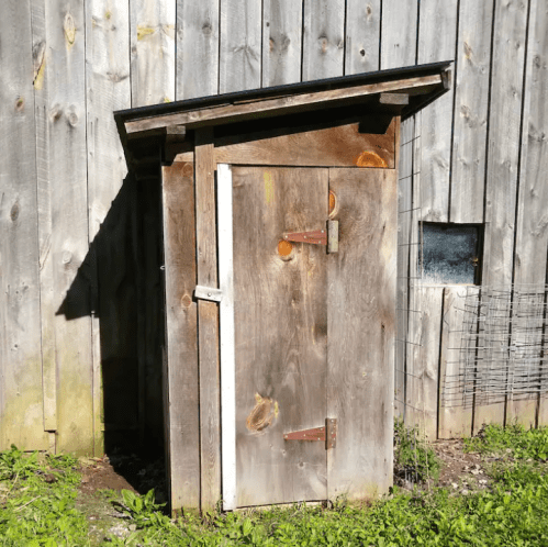 A small wooden shed with a slanted roof, standing against a weathered wooden wall, surrounded by grass.