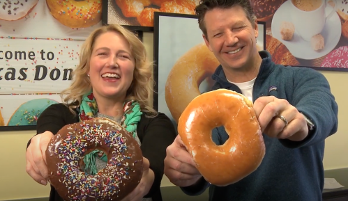 Two smiling people hold up large donuts, one with chocolate and sprinkles, the other a glazed donut.