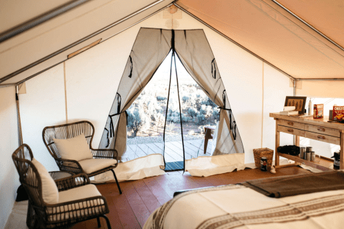 Interior of a glamping tent with a cozy seating area, wooden table, and open entrance revealing a natural landscape outside.