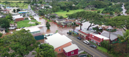 Aerial view of a flooded area with houses, trees, and a road, showcasing significant water accumulation.