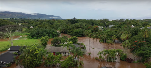 Aerial view of a flooded area with houses surrounded by water and lush greenery, under a cloudy sky.