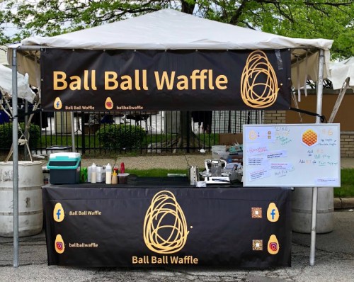 A food stall with a black banner reading "Ball Ball Waffle," featuring a menu and waffle-making equipment.