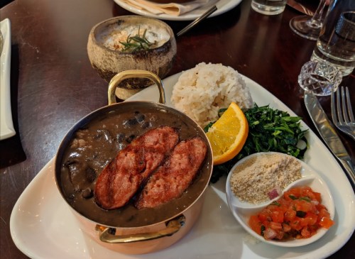 A plate featuring black beans with sausage, rice, sautéed greens, orange slice, and a side of salsa and seasoning.